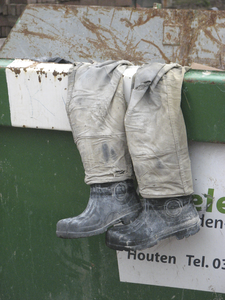 908026 Afbeelding van een weggegooide werkbroek met daarin twee rubber laarzen gestoken, over de rand van een container ...